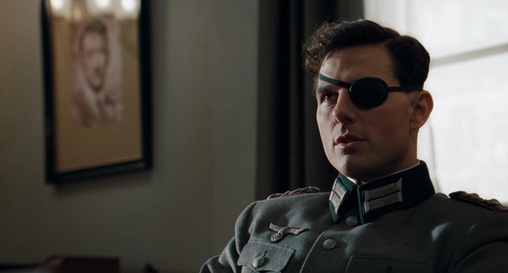 A man wearing an eye patch and gray uniform speaks to someone off camera. An out of focus portrait of Adolf Hitler is in the background