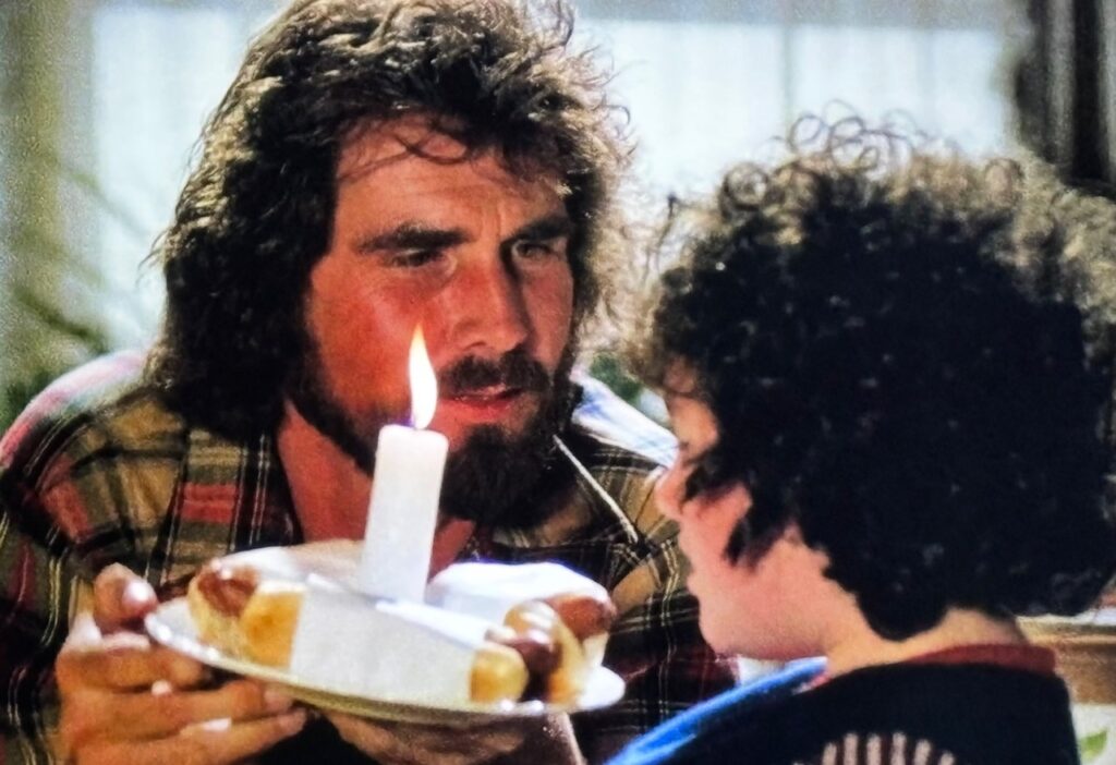 ames Brolin as Sean Boyd presents a plate of hot dogs with a lit candle center to his daughter, actor Abby Bluestone, in her bed on her birthday