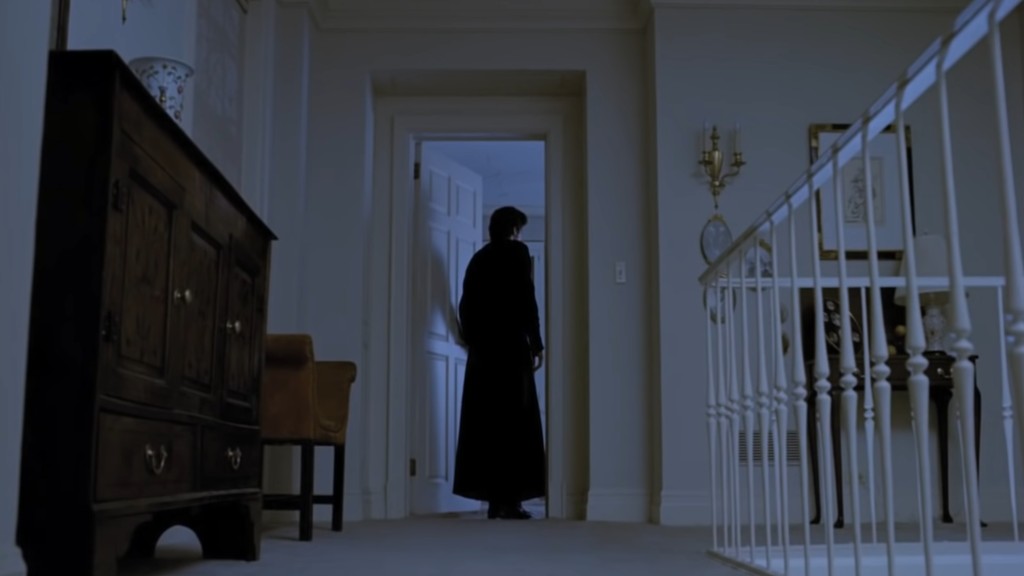 Father Karras is seen from behind as he stands in the doorway of Regan's room looking in. A blue light inside the room casts his shadow on the door.