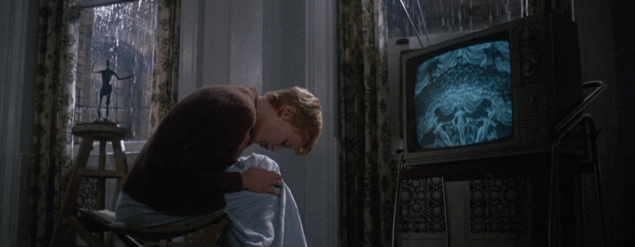 Rosemary, a light-skinned young woman with short blonde hair, cradles her knees bending forward while sitting on a stool in front of a television that shows a burlesque dance sequence. The TV glows blue, and the room she is in is draped in beige-patterned curtains, framing tall windows that show heavy rain outside.