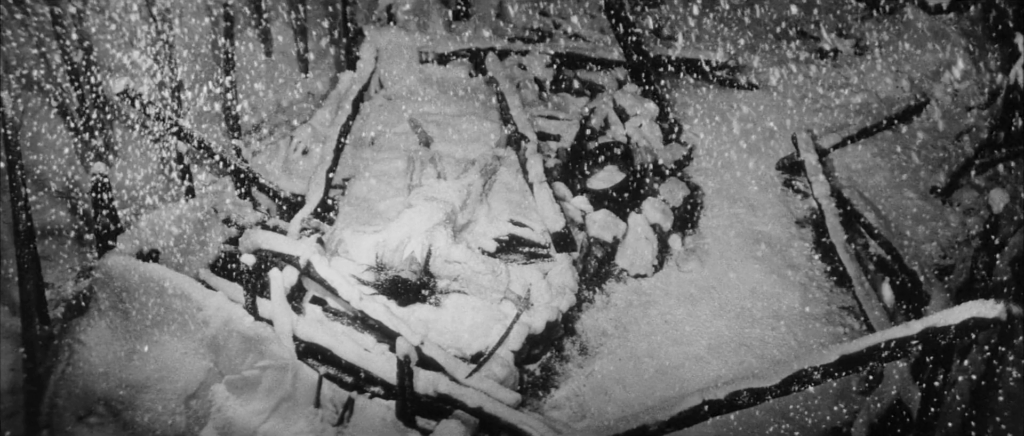 A dead man lies face up in the burnt remains of a house as it is snowing. A thin blanket of snow covers everything.