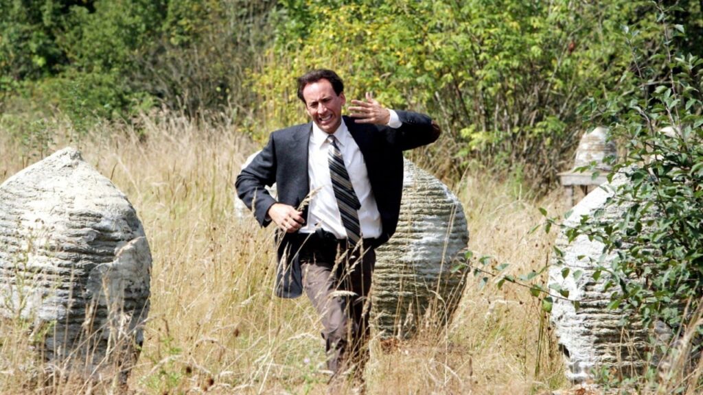 A distressed man in a dark suit runs through overgrown grass, his face twisted in pain as he clutches at the air. Surrounded by strange beehive-like stone structures, he appears trapped in a frantic moment of confusion and fear.