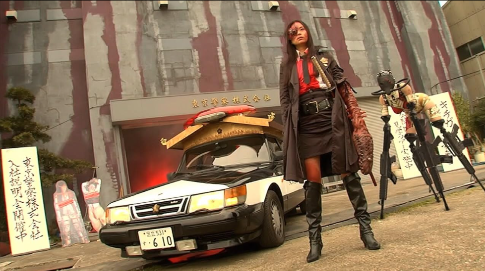 A woman in Black Trench coat with a snake head arm stands in front of a police car. Behind her is a woman in a gas mask with machine guns for arms and leg.