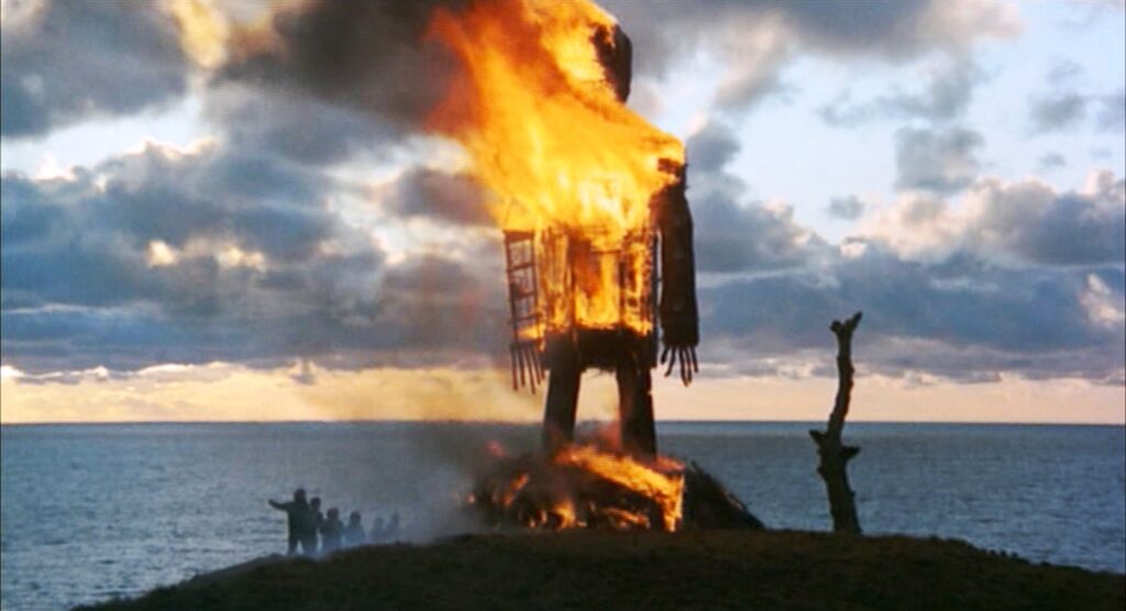 A large wicker figure shaped like a human burns against a dramatic cloudy sky at sunset, standing on a cliff overlooking the sea. Small silhouetted figures watch from a distance as flames consume the structure.