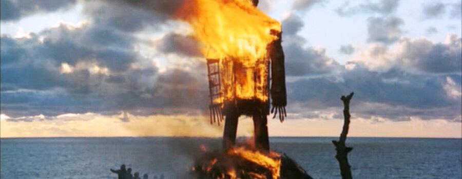 A large wicker figure shaped like a human burns against a dramatic cloudy sky at sunset, standing on a cliff overlooking the sea. Small silhouetted figures watch from a distance as flames consume the structure.