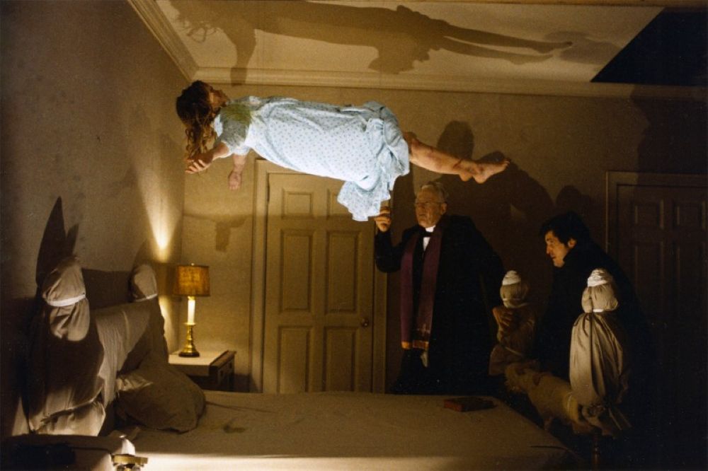 Linda Blair as Regan hovers over her bed in a light blue nightgown as Father Karras and Father Merrin pray next to the bed.