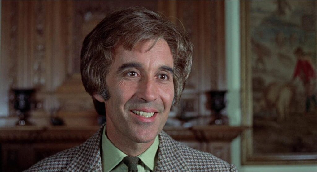 A man with graying hair and sideburns smiles warmly, wearing a green shirt, a green tie, and a checkered brown blazer. He stands in a richly decorated room with wood paneling and a large painting behind him, giving the scene an air of old-world refinement.