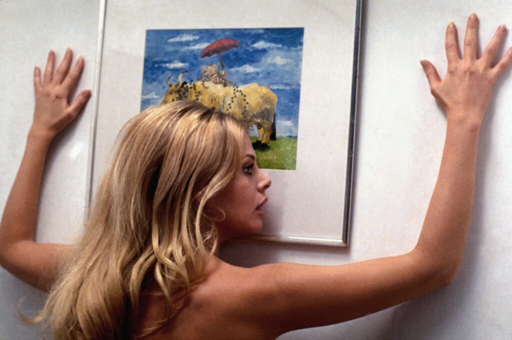 A blonde woman presses her body and hands against a white wall, gazing sideways at a framed painting of cows under a red umbrella in a grassy field. The scene is brightly lit, with the painting providing a colorful contrast to the stark wall.