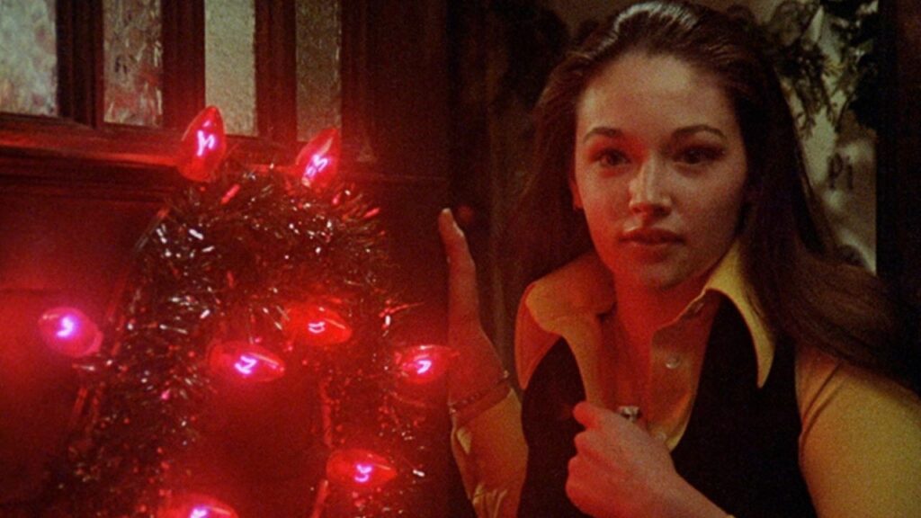 A young woman wearing a yellow, collared shirt and dark vest looks out a door decorated with a wreath and glowing red lights.