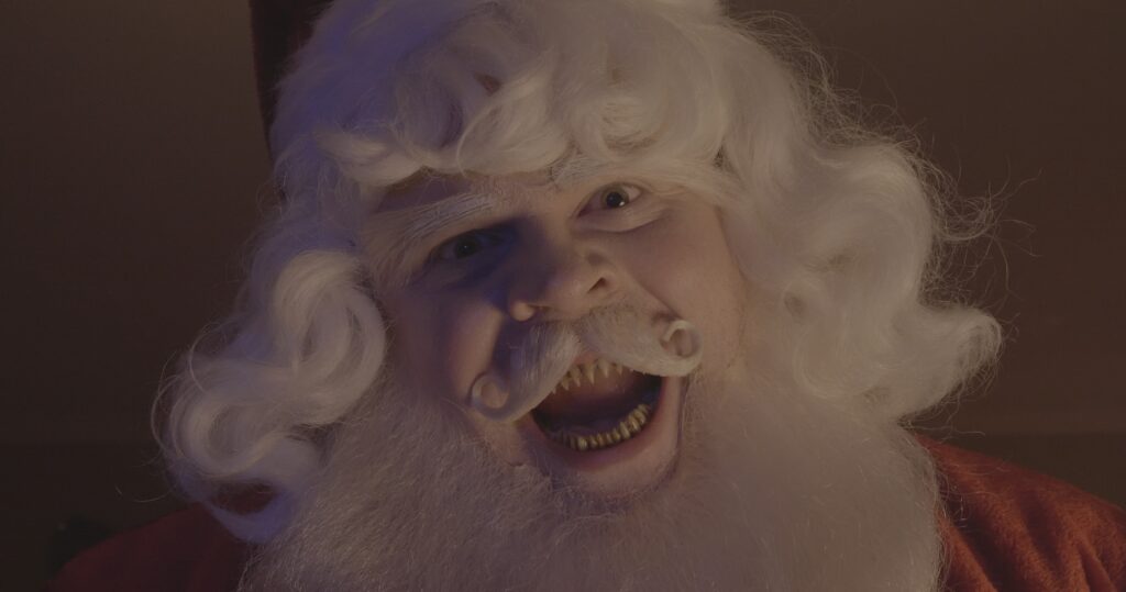A close up of Santa Claus looking directly into the lens. His mouth is open wide, revealing sharp teeth.