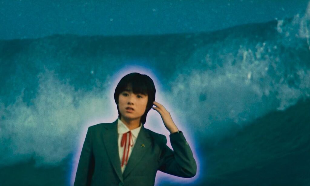 A Japanese woman, wearing a suit, stands in front of a crashing wave.