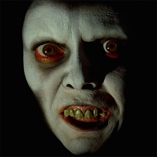 An up close shot of Pazuzu, the literal demon possessing Regan. He features bone white skin, red rimmed, brown eyes, gaunt cheeks, and yellowed, decayed teeth.