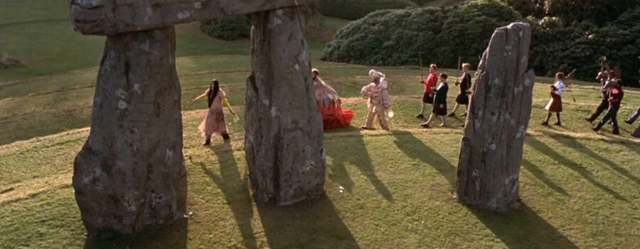 A procession of wildly dressed people weave through a Stonehenge-like structure.