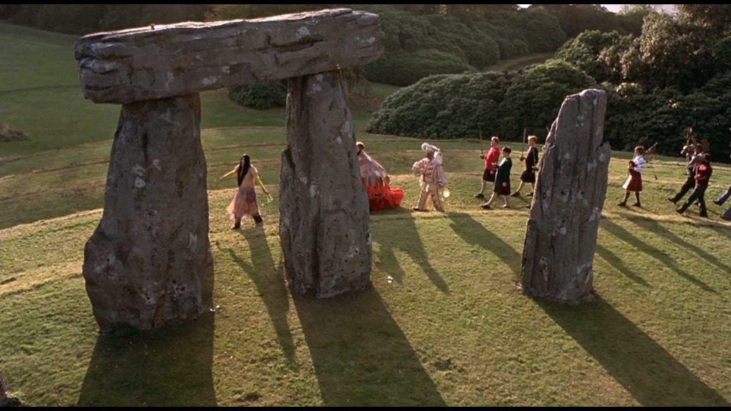 A procession of wildly dressed people weave through a Stonehenge-like structure.