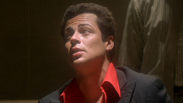 Benicio del Toro in a red shirt and black jacket, talking back to police in a dark room