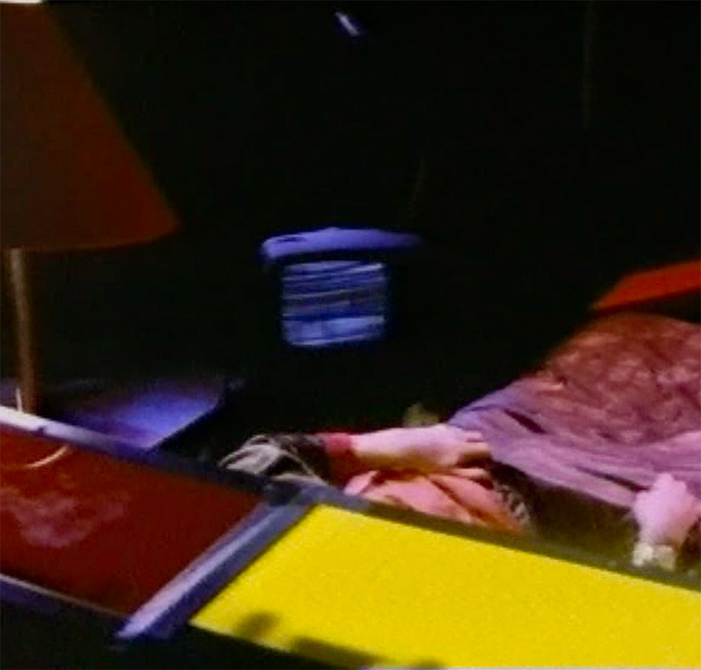A bedroom containing a small television covered with a shawl, which darkens the wavy black-and-white lines on the screen. In the foreground, Two arms and a torso are partially shown under covers in a bed made of rectangles, one red the other yellow.
