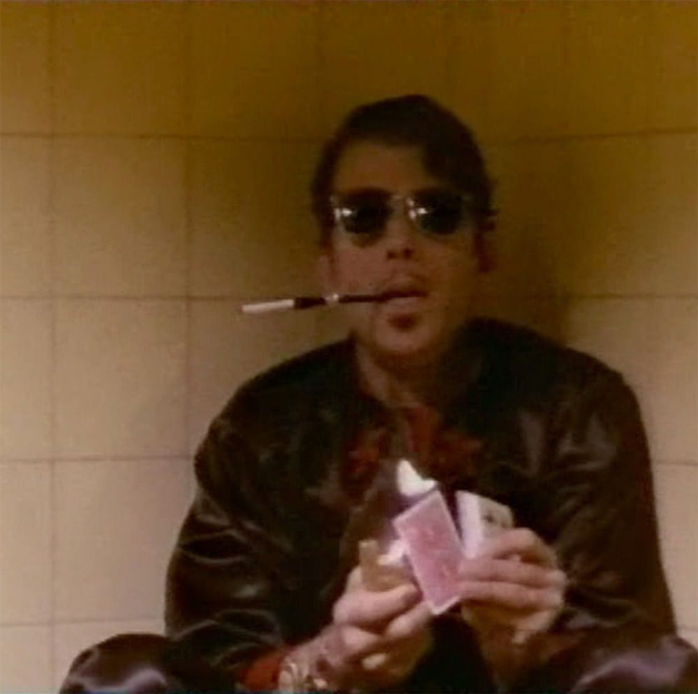 A man seated against a tiled corner uses a gold lighter to light a playing card. He wears sunglasses indoors. He has a long cigarette holder with cigarette in his mouth.