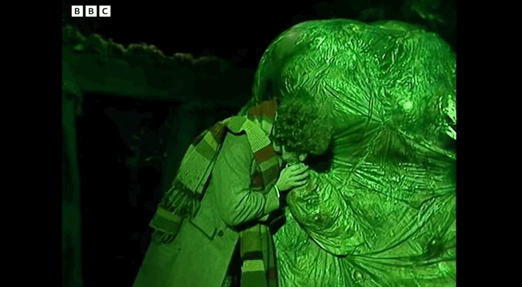 The Doctor stands by the creature, a giant green blob, and holds its appendage up to his mouth.