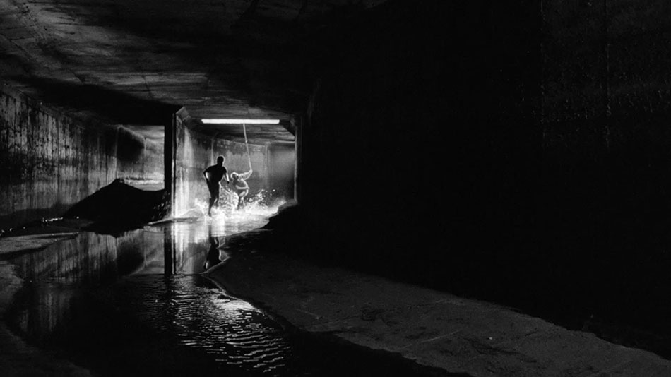 A dark sewer, lit brightly from a hole in the ceiling. A rope dangles from that hole. Bob (Roberto Benigni) splashes in the water as he slides down that rope. Zack (Tom Waits) runs ahead towards the camera.