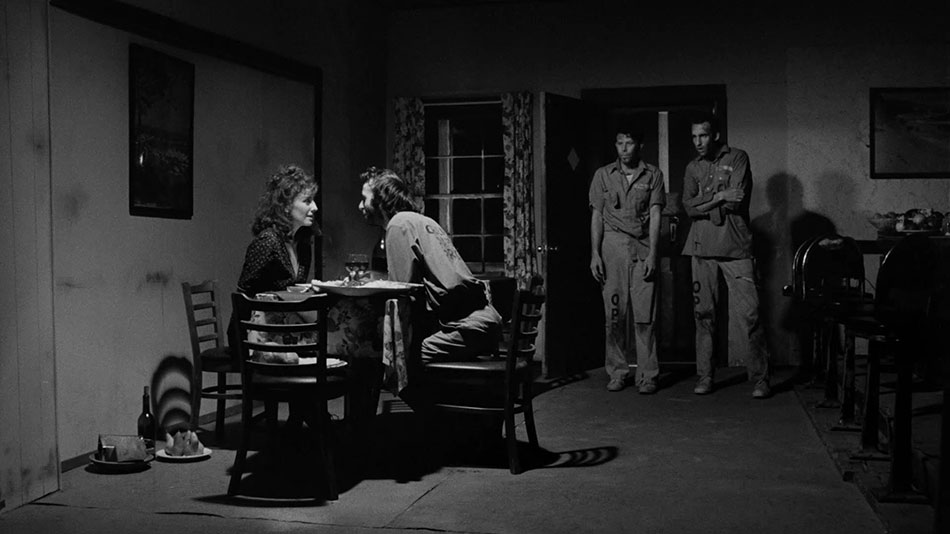 Nicoletta (Nicoletta Brasci) and Bob (Roberto Benigni) lean toward one another affectionately at a round dinner table in Nicoletta’s kitchen. The two are brightly lit. Zack (Tom Waits) and Jack (Jon Lurie) stand in the shadows by the doorway looking confused, amused, and a bit ragged. Bob, Jack, and Zack are all wearing matching prison jumpsuits.