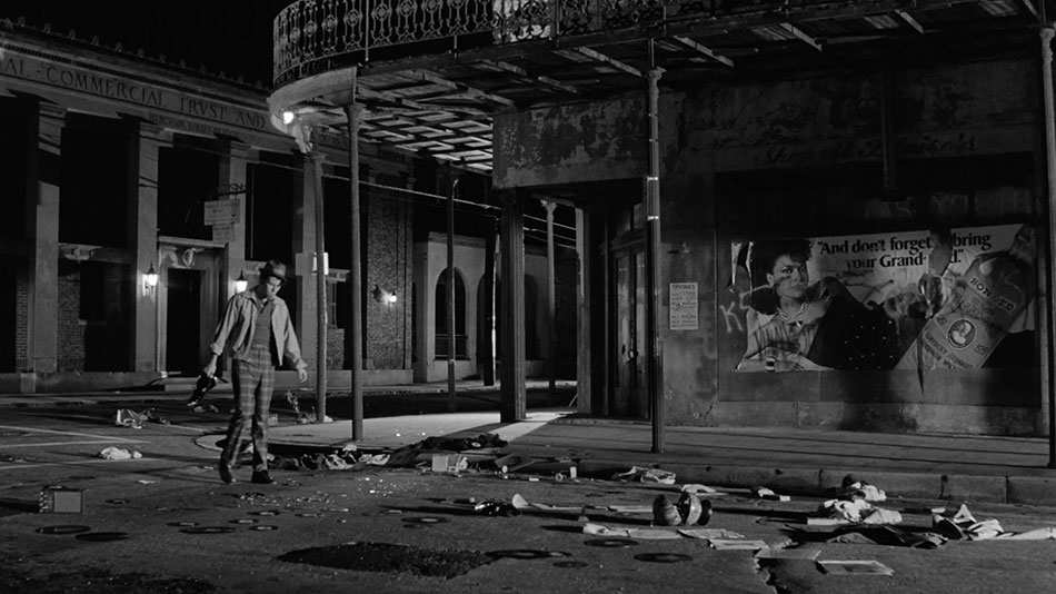 Zack (Tom Waits) in tweed pants, light jacket, and hat stands amid the wreckage of his belongings shattered along the sidewalk of this dimly lit New Orleans street at night. To his right sits a defaced poster of a woman holding a bottle of some kind of alcohol. Part of the poster has been torn off and is therefore illegible.
