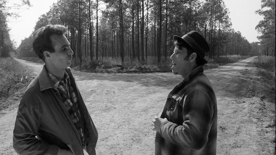 Left to right, Jack (John Lurie), wearing a leather jacket and checkered shirt, and Zack (Tom Waits) in a flannel coat and hat, stand at a fork in the road facing each other. Trees line the edge of the road and the sky is bright.
