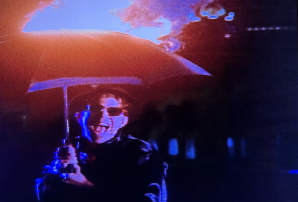 Tom Waits, wearing sunglasses and a porkpie hat, laughs maniacally while holding an umbrella on fire.