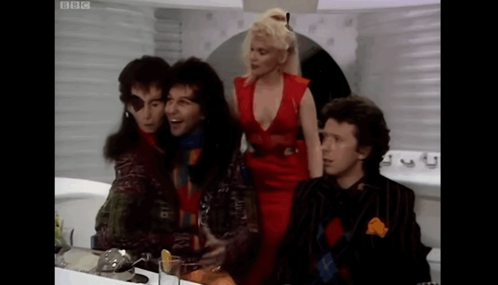 Zaphod Beeblebrox and Ford Prefect are seated together with Trillian standing nearby. Zaphod’s second head droops glumly.