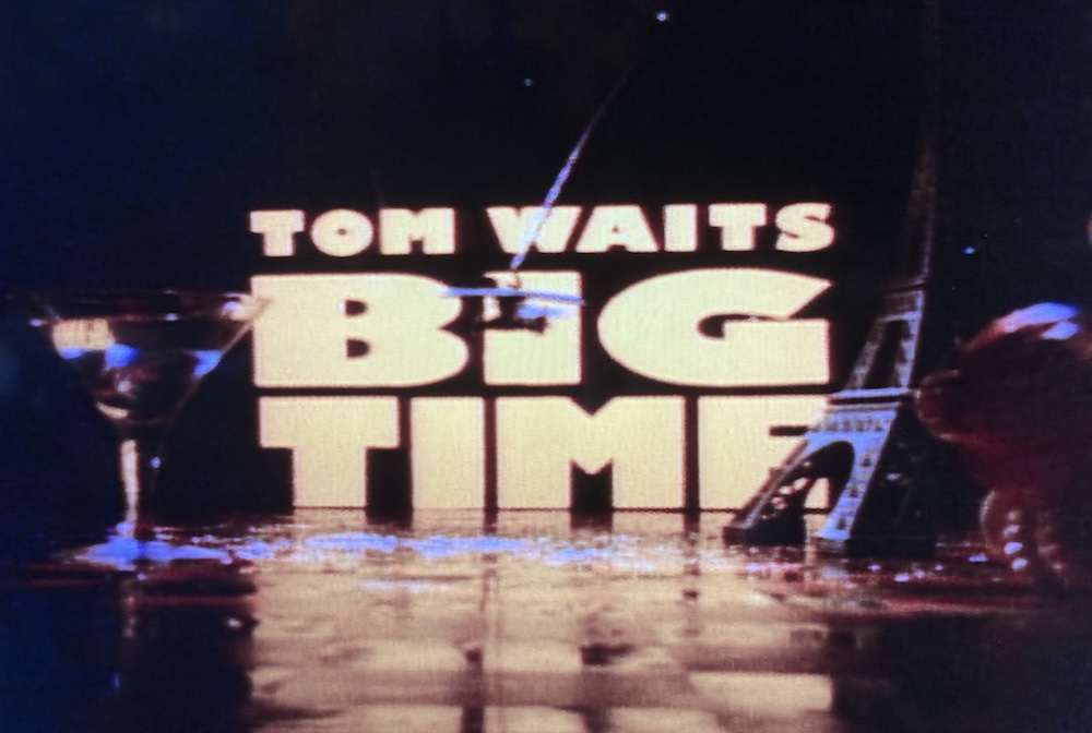 Title card to the concert film “Tom Waits: Big Time” featuring large yellow script, with Martini glass and miniature Eiffel Tower in the foreground.