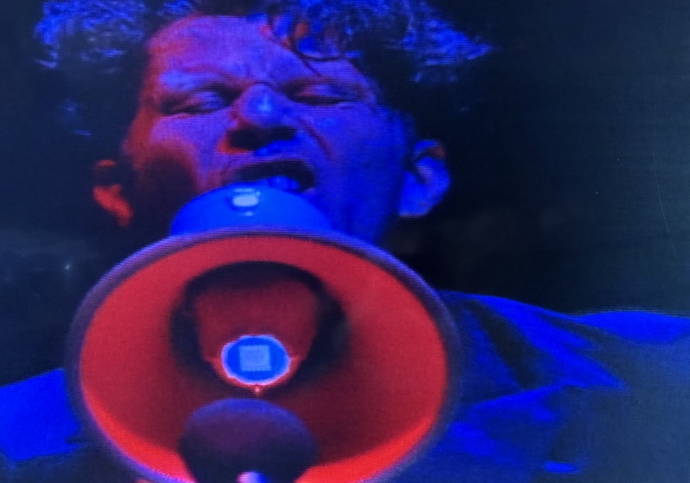 Tom Waits performing live, snarling into a bright red megaphone.
