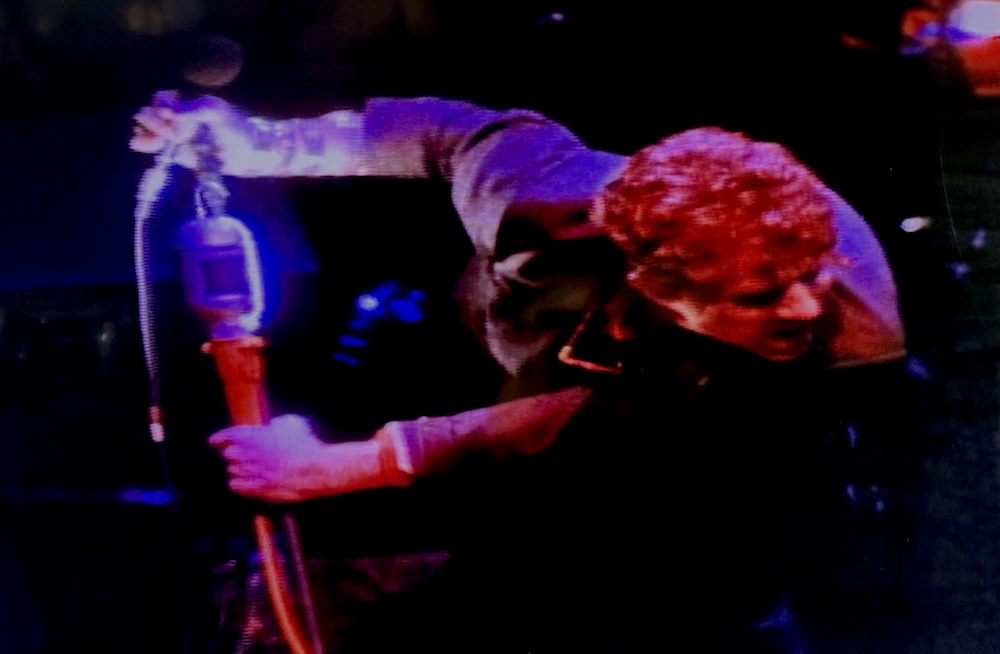 Tom Waits performing live, leans away from a microphone stand on which is mounted a construction light and bulb.
