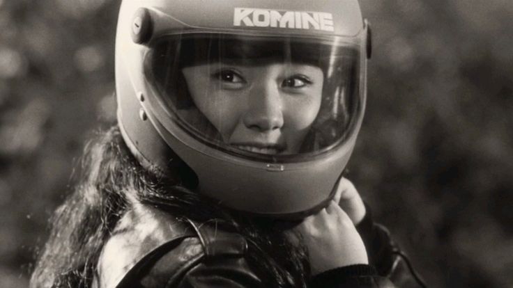 A still from the film of Miyoko in close-up, smiling, donning a black biker helmet and leather jacket.