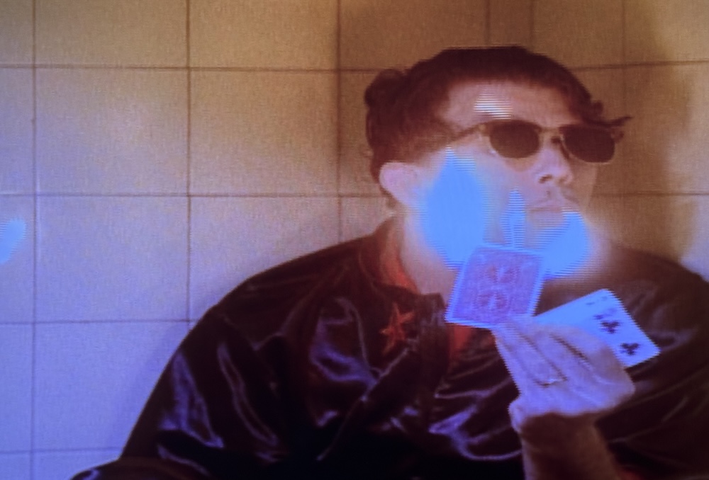 Tom Waits, dressed as Frank O’brien, sits on the floor of a public bathroom, lighting a cigarette with flaming playing cards.