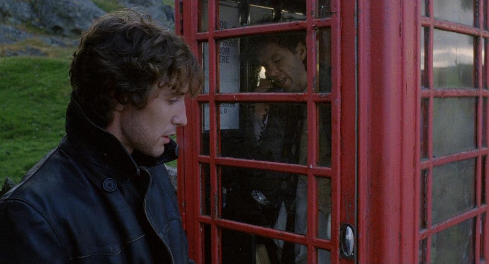 A man with curly brown hair and a black leather jacket stands on the leftside foreground looking down, with green grass and the bottom of a hill peeking out in the distance behind him. He's facing in profile towards the outside of a red phone booth, which fills the rest of the frame, its corner jutting out towards the viewer. Through the phonebooth's window there is a man with black hair speaking into its black receiver, and also looking down. The hills are reflected through the paned windows on the phonebooth's opposite side