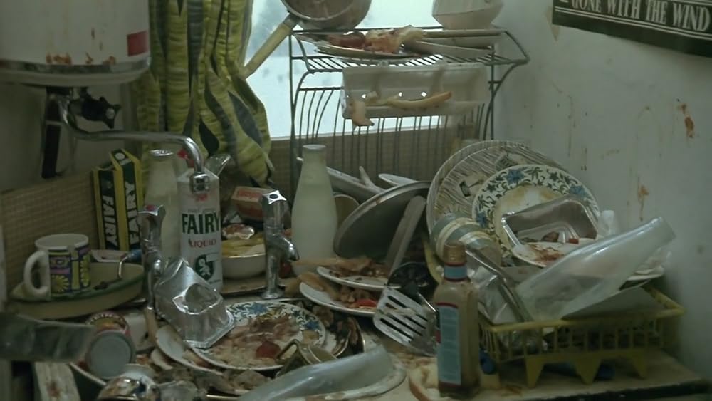 A tight shot on a kitchen sink overflowing with dirty dishes and empty bottles with a dish drainer beside it also filled with dirty dishes. Another dish with food on it appears to be piled on a higher rack and an opaque window sits right behind it with its curtain pushed to one side.