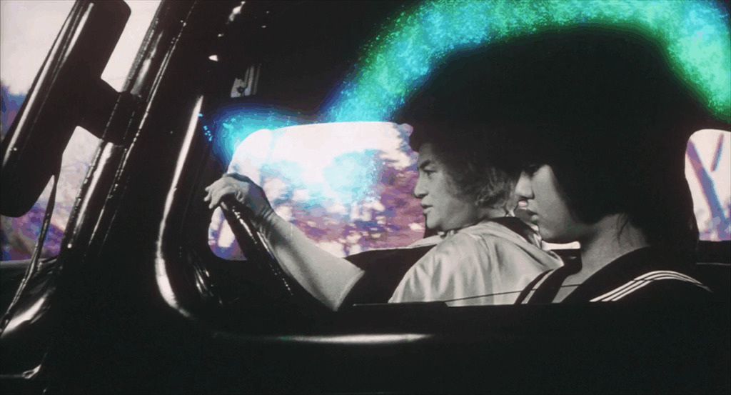 A side profile two-shot of Yuka (played by Hiroko Yakushimaru) and the demon Kyogoku (played by Tôru Minegishi) in the front half of a car, with Yuka in the foreground, head turned slightly down and to the right, and Kyogoku with hands on the steering wheel in the middle ground, looking resolute and straight ahead. The two figures are superimposed black-and-white on a color background of trees whizzing by through the vehicle's windows with a spotty sheen of neon green bouncing over the enemies' heads and Kyogoku's long-sleeved gloves.