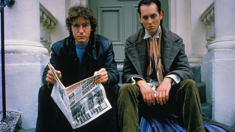 Two men sit on the front stoop in front of a closed doorway, facing the camera. 

The man on the left has curly brown hair and round John Lennon glasses. 

He wears a black leather jacket with a blue shirt underneath and he’s holding an open newspaper with a headline that reads "The Secret Hideout" but the rest of the headline is obscured. He’s looking into the camera. The man to his right is making a sullen face and has black slicked back hair. He’s wearing an overcoat, a shirt with a white collar and a pink and gray scarf. He’s wearing olive green trousers and sits with his knees apart.