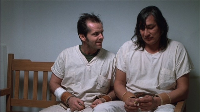 An excited and enticed Randle Patrick McMurphy is looking at Chief Bromden, both men sit on a wooden bench