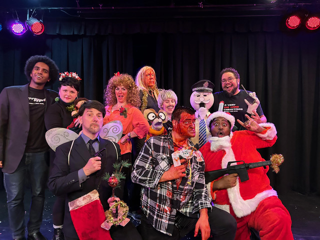 A group of performers pose together on a small stage, many wearing Christmas-themed costumes and holding props.