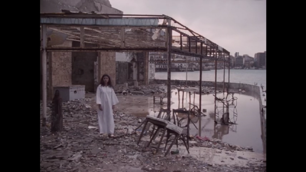 A woman in a white tunic dress stands on the dilapidated patio of a bayside restaurant, a ruined city in the background.

