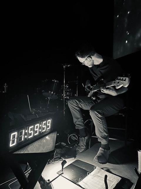 On the left side of this black-and-white shot is a Hartke amp, branded white letters bold against the blackness. On the right side is PRGRPHS guitarist and synth player Chris Polley, tapping the fretboard with an array of pedals at their feet. A reading light atop the synths is overexposed, flaring across the frame.