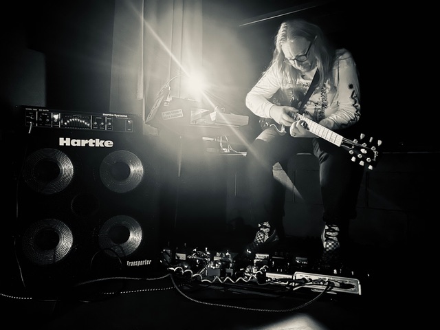 On the left side of this black-and-white shot is a Hartke amp, branded white letters bold against the blackness. On the right side is PRGRPHS guitarist and synth player Chris Polley, tapping the fretboard with an array of pedals at their feet. A reading light atop the synths is overexposed, flaring across the frame.
