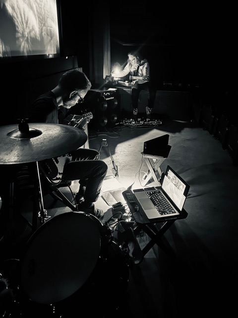 A long shot from behind the drum kit with cymbals in the foreground, Conway in the middle ground, and Polley in the background. The Trylon screen peeks into frame from the left side and a MacBook sits to the right on a stand.