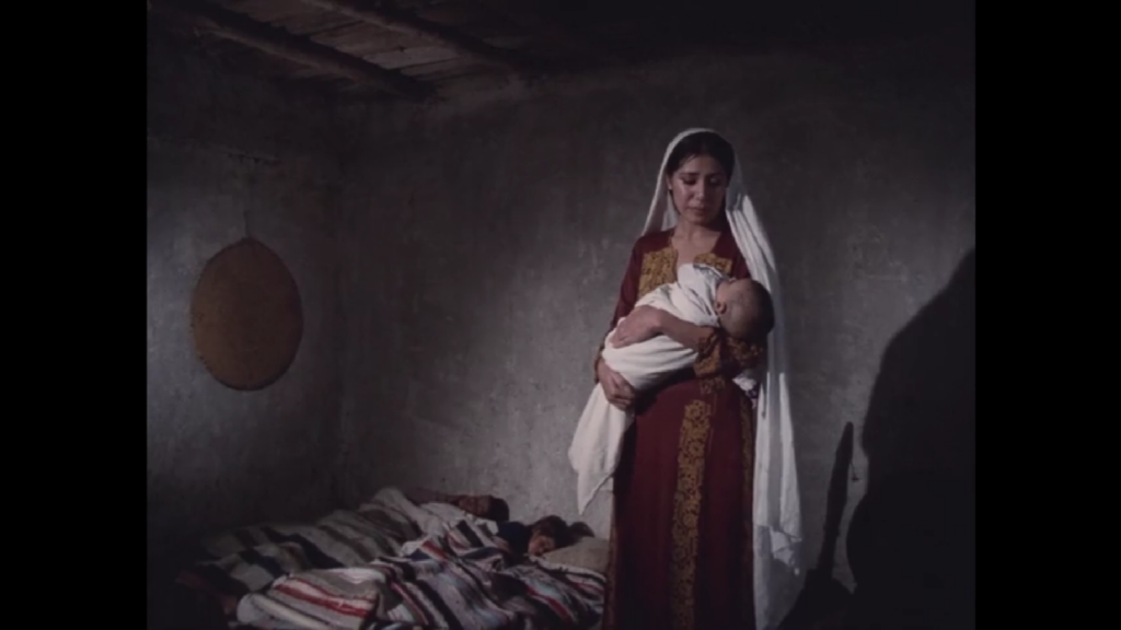 In a dimly lit, barren-walled room, two children sleep on a bed on the floor, while a mother stands holding her infant.