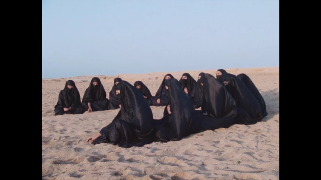 13 women in black burqas sit in a semi-circle on the beach, uncomfortable, facing the sea where their husbands and sons play.