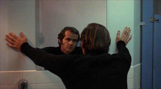 A man looks at his reflection in a mirror that he straddles with his arms.