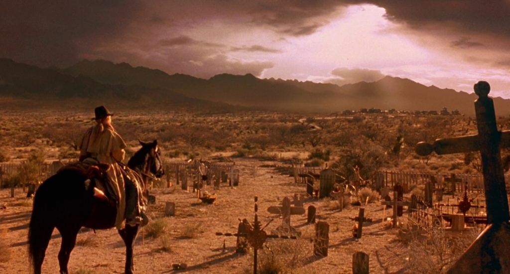 A sepia-toned frame. Ellen “The Lady” McKenzie (Sharon Stone) in poncho and cowboy hat looks at mountains in the horizon from atop her horse, positioned in the middle of a graveyard.
