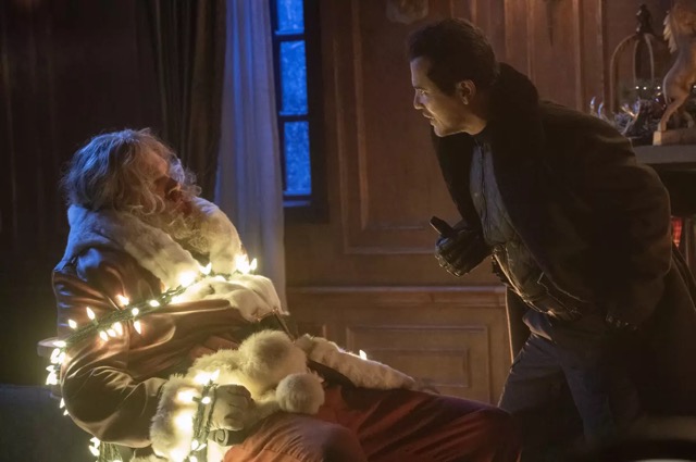 A man in dark clothing leans toward a seated Santa Claus who is bound to a chair with glowing Christmas lights wrapped around him