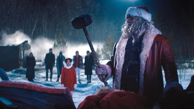 Santa Claus stands in a snowy outdoor setting at night holding a large hammer. Several people stand behind him watching, including a young girl in a red coat.