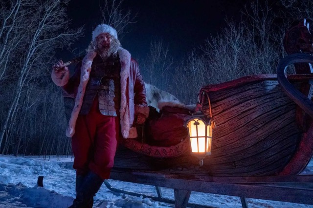 Santa Claus stands beside a wooden sleigh in a snowy forest at night, holding a sledgehammer. His face and coat are smeared with blood, and a lantern glows from the side of the sleigh.